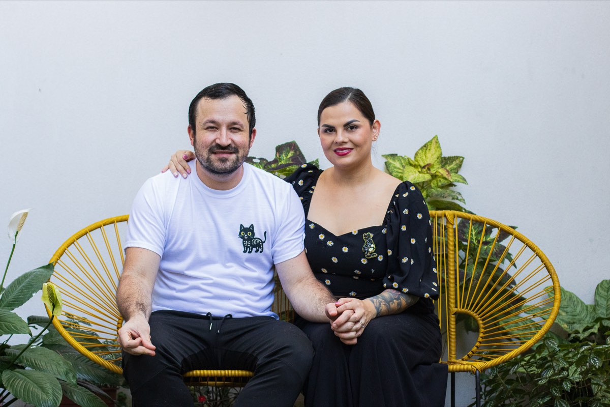 Ari y Tony, la pareja detrás de Salem House, sentados en una banca de mimbre amarilla rodeados de plantas.